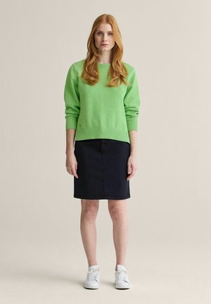 Kvinde med langt rødt hår iført en lys grøn sweater, sort nederdel til knæet og hvide sneakers, stående mod en ensartet baggrund.