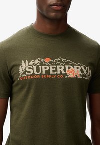 Olijf groene katoenen t-shirt met een afbeelding van bergen en bos, "SUPERDRY" logo in het wit, en oranje accenten. Korte mouwen en ronde hals.