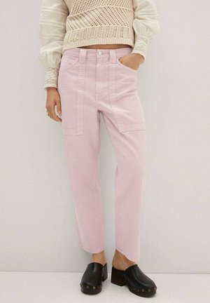 Persoon draagt lichtroze broek met hoge taille en opgestikte zakken, crèmekleurige gebreide trui met pofmouwen, en zwarte instapklompen met studs.