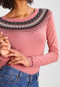 Pull rose à manches longues avec un empiècement brodé noir et blanc présentant des motifs géométriques, associé à un jean en denim bleu.