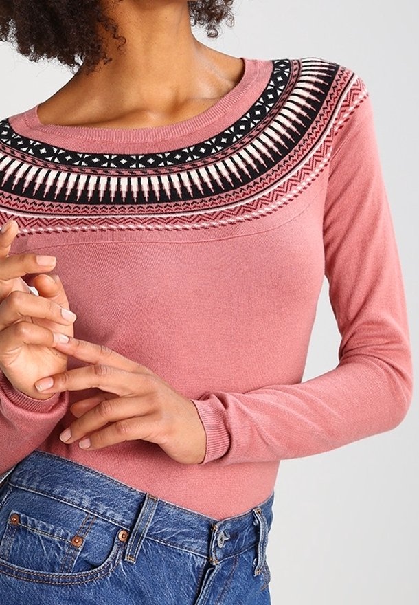 Pull rose à manches longues avec un empiècement brodé noir et blanc présentant des motifs géométriques, associé à un jean en denim bleu.