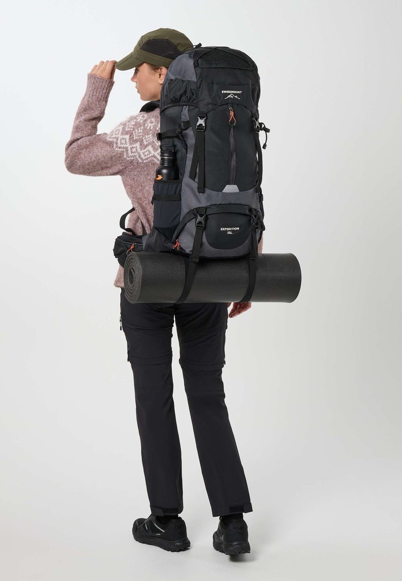 Personne portant un grand sac à dos de randonnée noir et gris avec un tapis de sol roulé attaché, vêtue de vêtements de plein air et une casquette verte.