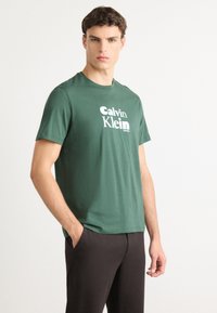 Man wearing green Calvin Klein t-shirt and black pants, standing with one hand in pocket against plain white background.