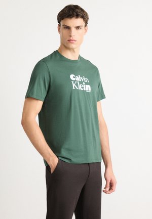 Man wearing green Calvin Klein t-shirt and black pants, standing with one hand in pocket against plain white background.
