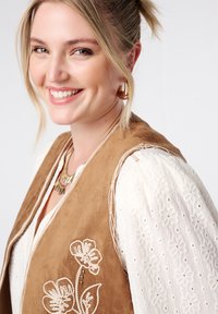 Bruine suède vest met bloemborduurwerk, gelaagd over een witte, textuurrijke blouse. Gouden sieraden accentueren de outfit.