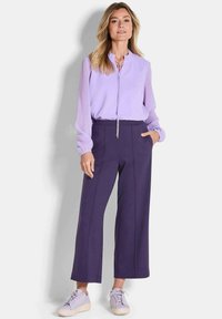 Lilafarbene Bluse mit gerafftem Ausschnitt und Bund, kombiniert mit hochgeschnittenen, weiten, dunkelpurpurnen Hosen und hellpurpurnen Turnschuhen.