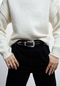 Ceinture noire avec ferrures argentées et clous décoratifs, portée sur un pull crème et un pantalon en velours côtelé noir ; bagues sur la main exposée.