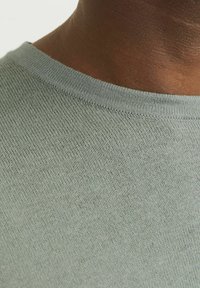 Pulover tricotat de culoare verde deschis, cu decolteu rotund. Materialul are o textură fină și un aspect neted, evidențiind detaliile cusăturii.