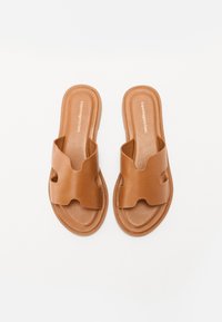 Braune Slipper aus glattem synthetischem Material, mit einem wellenförmigen Riemendesign und einer flachen Sohle. Minimale Details auf dem Fußbett.
