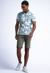 Vaaleansininen poolopaita kukkakuvioilla, yhdistetty oliivinvihreisiin, käännettyihin shortseihin. Malli käyttää valkoisia lenkkitossuja, esitellen rentoa kesäasua.