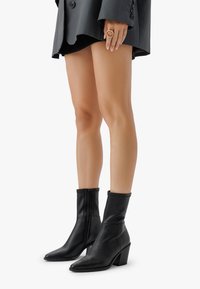 Botas negras hasta el tobillo hechas de cuero texturizado, con punta afilada y tacón cuadrado, y cierre de cremallera en la parte posterior.