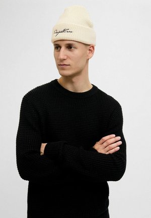 Ung mand iført en cremefarvet strikket hue med sort tekst og en sort struktureret sweater, armene krydset, kigger til siden mod en ensfarvet baggrund.