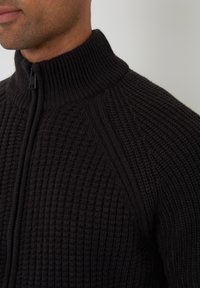 Mörk, ribbad stickad tröja med hög krage och dragkedja, med texturerat tyg och raglansägen.