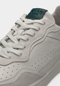 Κοντινό πλάνο λευκού δερμάτινου αθλητικού παπουτσιού με σουέτ λεπτομέρειες, λευκά κορδόνια, διάτρητες πλευρικές λεπτομέρειες και μαύρο λογότυπο στο γλώσσα.