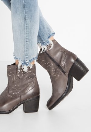 Stivaletti grigi in pelle alla caviglia con borchie decorative e cerniere laterali, indossati con jeans blu chiaro con orlo sfrangiato.