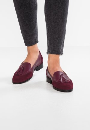Voeten met donkerrode leren en suède loafers met kwastjes, gecombineerd met zwarte skinny jeans met afgewerkte zoom op een witte ondergrond.