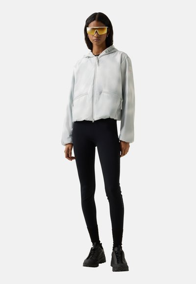 Vrouw die een oversized lichtgrijze zip-up hoodie draagt, zwarte legging, zwarte chunky sneakers en reflecterende oversized zonnebril staand.