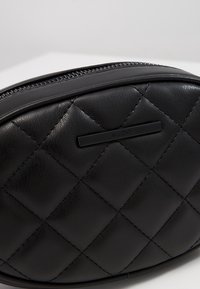 Bolso negro de cuero sintético acolchado con forma redondeada. Presenta un cierre superior con cremallera y un acento de logotipo negro mate. Superficie texturizada con patrón de diamantes.