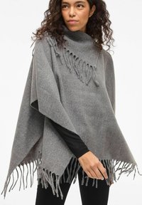 Sieviete, kas valkā pelēku pončo ar svārkiem pār melnu garu piedurkņu topu un melnām biksēm, stāv uz viendabīga gaiša fona.