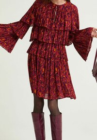 Robe rouge à motif floral avec des volants superposés, des manches longues en cloche et un col en trou de serrure. Portée avec des bottes bordeaux montantes jusqu'aux genoux.