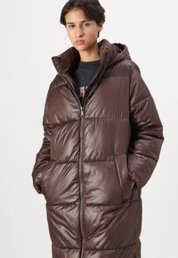Abrigo tipo puffer marrón con capucha, que presenta un cierre con cremallera en la parte delantera, diseño acolchado y dos bolsillos laterales. Textura brillante con un corte de longitud media.