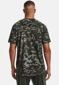 Zelená kamuflážní košile s krátkým rukávem, která obsahuje směs tmavých a světlých vzorů, vyrobená z lehkého materiálu s kulatým výstřihem.