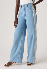 Person wearing light blue wide-leg jeans with silver side stripes and beige open-toe heels, standing against a plain background.