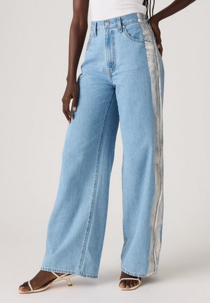 Person wearing light blue wide-leg jeans with silver side stripes and beige open-toe heels, standing against a plain background.