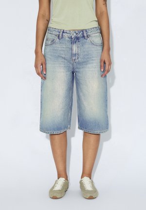 Persona que lleva puestos unos pantalones cortos vaqueros de mezclilla azul claro hasta la rodilla, una camiseta sin mangas de color amarillo pálido y zapatillas verdes con textura y cordones blancos, de pie.