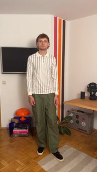 Pikend varrukatega, kreemikas valge polo särk, millel on vertikaalsed mustad triibud; oliivrohelised cargo püksid; mustad libisevad kingad; seisab puidust põrandal.