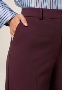Bordeauxkleurige broek met een gladde textuur, voorzien van zijzakken en een aansluitende tailleband, gecombineerd met een lichtblauw gestreept shirt.
