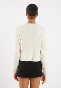 Pulover din tricot ribatat crem, cu mâneci lungi, lungime cropped și decolteu rotund, asortat cu o fustă scurtă neagră. Textură netedă.
