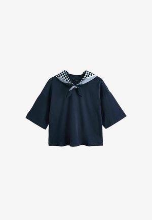 Tengerészkék rövid ujjú ing széles ujjakkal és fekete-fehér pöttyös sállal, amely a nyaknál van megkötve.