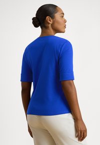 Žena so zopnutými vlasmi nosiaca kráľovskomodré tričko s krátkymi rukávmi a svetlobéžové nohavice, stojaca z profilu na jednoduchom pozadí.