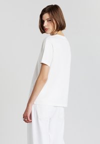 Giovane con capelli corti castani che indossa una camicia bianca a maniche corte e pantaloni bianchi, mentre si guarda oltre la spalla su uno sfondo chiaro e uniforme.