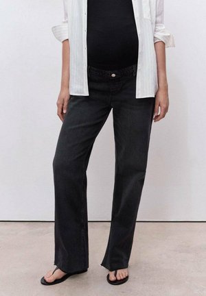 Pregnant person wearing black maternity jeans, black top, white open shirt, and black sandals, standing against a plain wall.