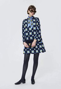 Kobieta w granatowej sukience z dużymi jasnoniebieskimi i żółtymi wzorami kwiatowymi, w czarnych rajstopach, czarnych butach, w okularach przeciwsłonecznych, trzymająca czarną kopertówkę.