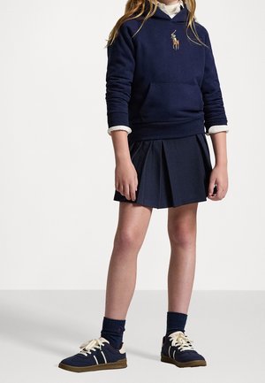 Barn iført navyblå hættetrøje med polo-logo, hvid skjorte, navyblå plisseret nederdel, navyblå sokker og navyblå sneakers med hvide snørebånd, stående.