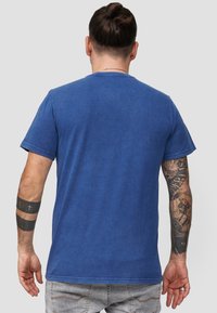 Sininen puuvillapaita, jossa on pyöreä kaula-aukko, lyhyet hihat ja hieman haalistunut tekstuuri. Näyttää takanäkymän, jossa on tatuointeja vasemmassa kädessä.