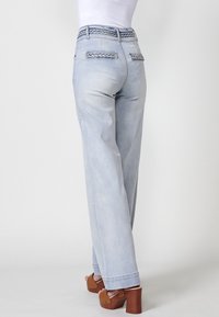 Jean taille haute bleu clair avec détails tressés à la taille et aux poches, coupe ample et légère déchirure. Associé à des sandales à plateforme marron.