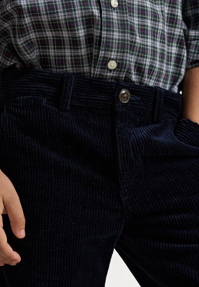 Pantalones de pana azul marino con acanalado vertical, equipados con un cierre de botón en la parte frontal, llevados con una camisa a cuadros verde y blanca.