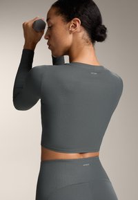 Szürke bordázott hosszú ujjú crop top, kerek nyakkivágással. Illeszkedő kialakítása van, és a hátán található a márkázott részlet, összeillő leggingszel viselve.