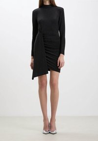 Kvinna i en svart långärmad minikjol med asymmetrisk drapering och silverfärgade spetsiga klackskor, stående på en ljus golvyta.