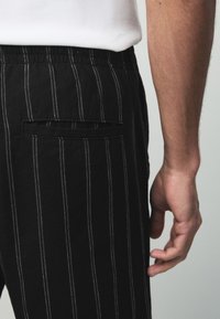 Schwarze gestreifte Hose mit elastischem Bund und einer Gesäßtasche. Der Stoff hat eine weiche Textur und kontrastierende weiße Nadelstreifen.