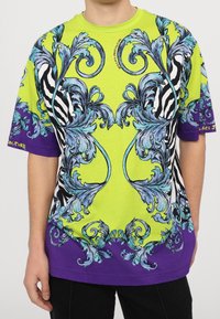 Tričko s krátkým rukávem v jasně zelené barvě, zdobené složitými modrými a černými vzory, s fialovým lemem a akcentem loga na rukávu.