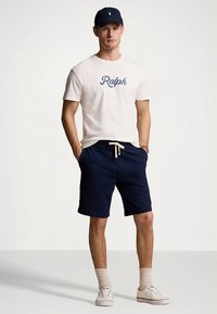Bílé bavlněné tričko s modrým nápisem "Ralph", doplněné temně modrými šortkami, krémovými teniskami a temně modrou čepicí. Neformální střih, minimalistický design, texturovaná látka.