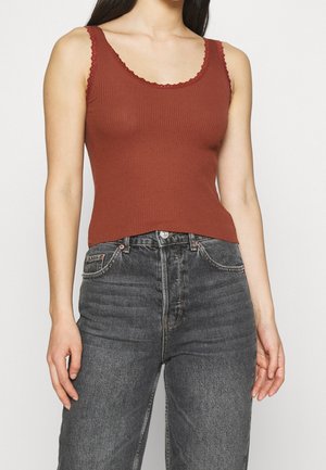 Débardeur côtelé couleur rouille avec bords ondulés, associé à un jean en denim foncé taille haute doté de poches avant et d'une braguette à boutons classique.