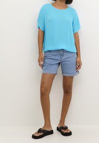 Personne portant un haut bleu ample à manches courtes, un short en jean retroussé, des sandales compensées noires, et un petit tatouage étoile sur le genou gauche.