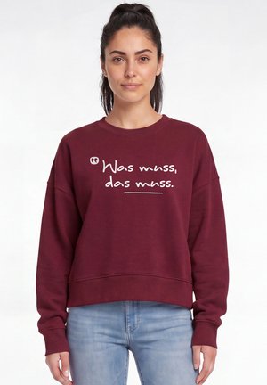 Frau mit dunklem Haar im Pferdeschwanz, die einen dunkelroten Pullover mit der Aufschrift "Was muss, das muss" und hellblaue Jeans vor weißem Hintergrund trägt.