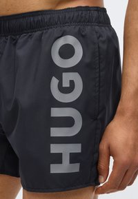 Melni peldkostīmi no viegla materiāla ar lielu pelēku "HUGO" logo sānos. Elastīga josta ar gludu tekstūru.